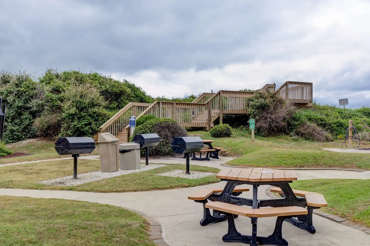 An outdoor grilling area complete with picnic table, barbecue grills, staircase, and a recycling bin, inviting guests to enjoy communal outdoor meals.