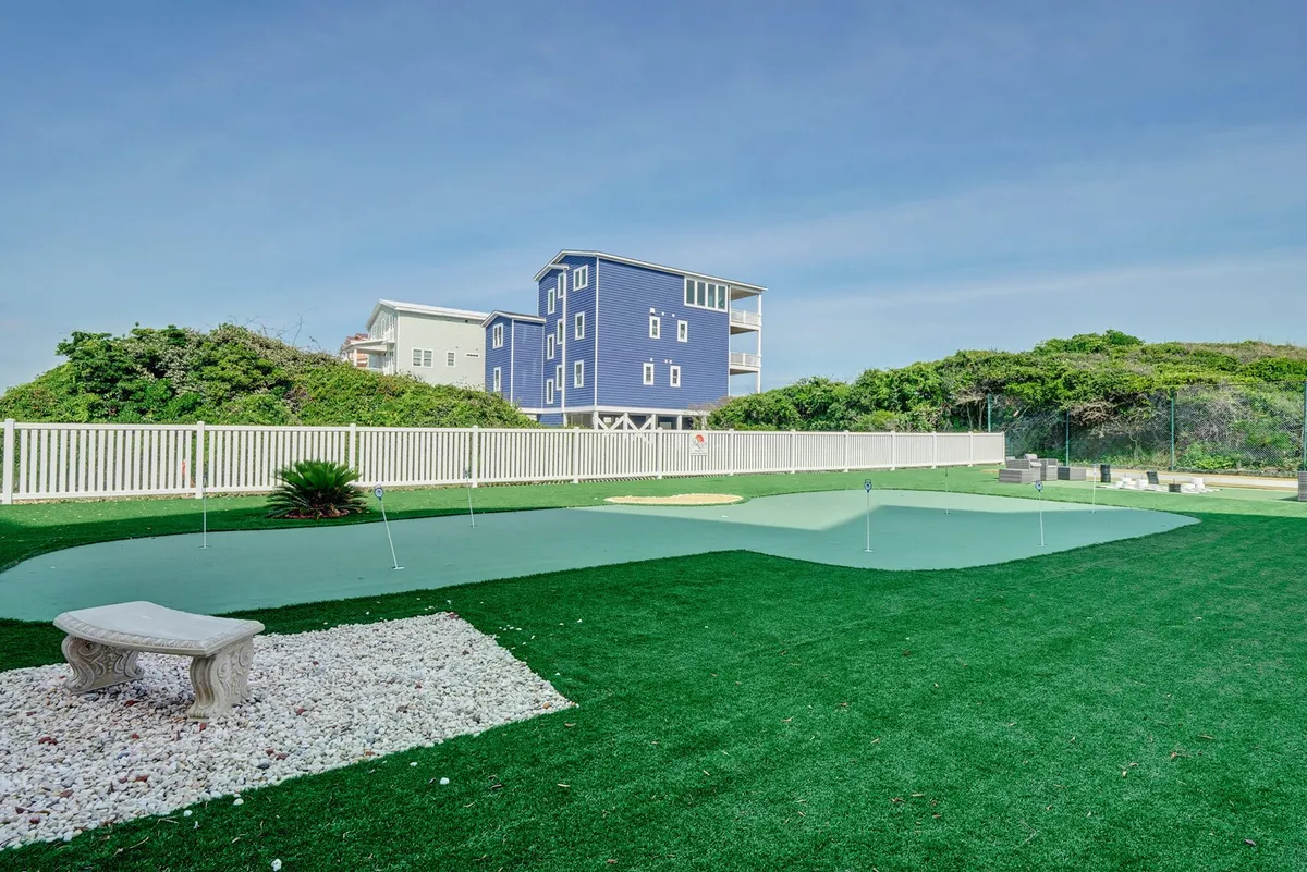 An exterior view of a putting green and a green lawn area, framed by a blue house and white fencing. The space is inviting for outdoor relaxation, casual sports, and family gatherings, emphasizing leisure and tranquility.