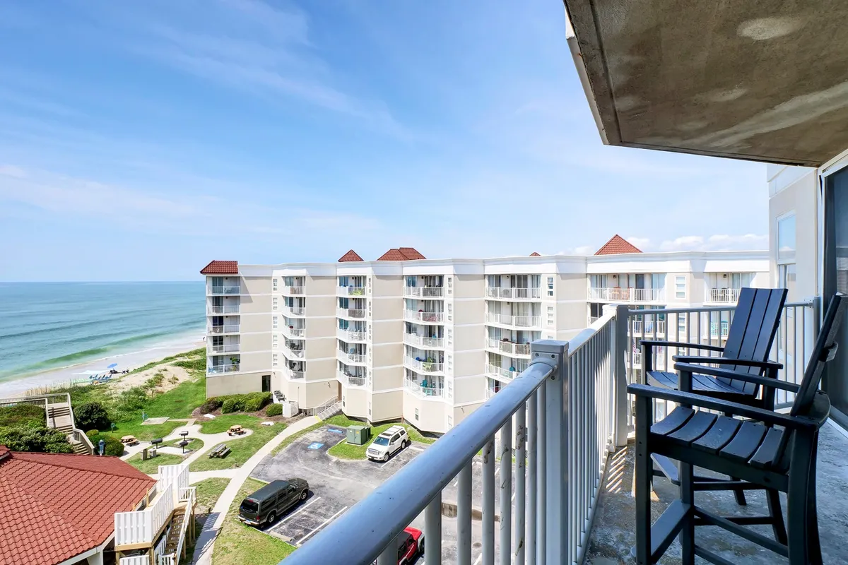 An inviting balcony with comfortable seating overlooking a sweeping ocean view, with the shoreline, sandy beach, and resort buildings in the distance. The tranquil setting is perfect for relaxing with friends or enjoying meals outdoors while feeling the sea breeze.