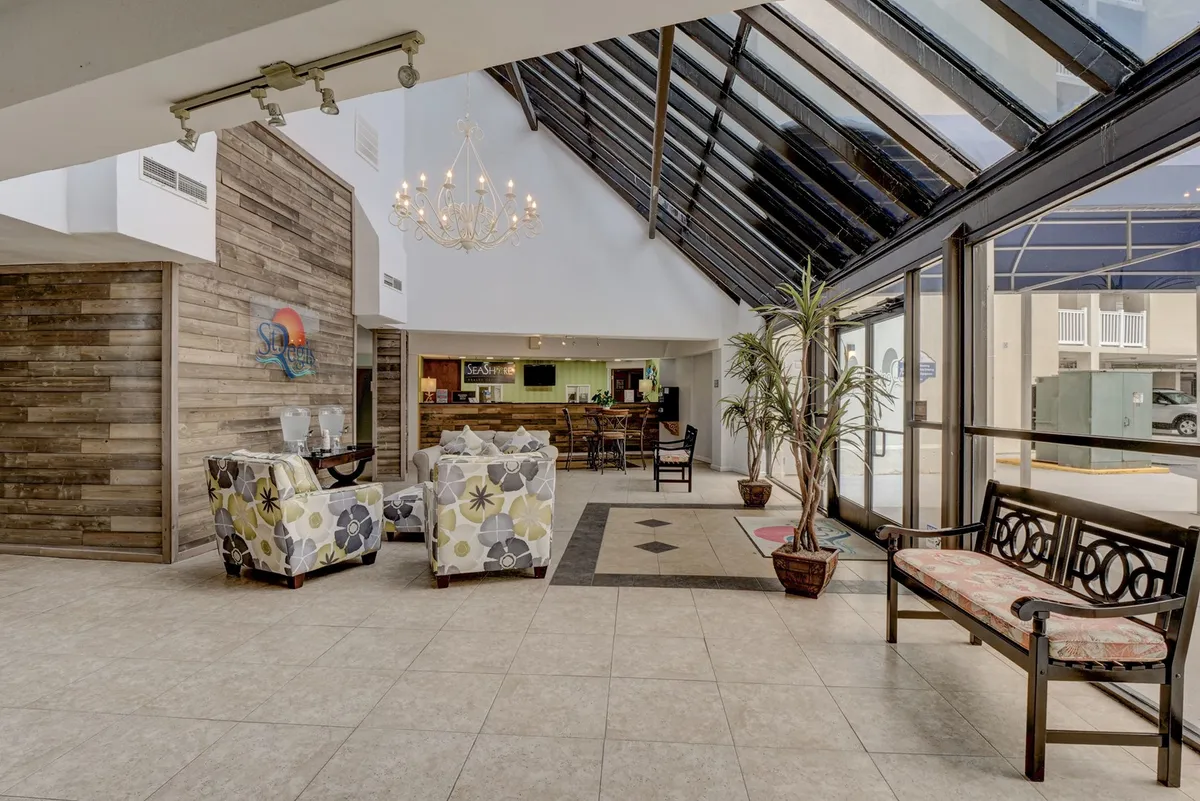 The image captures a welcoming resort interior featuring a chandelier, front desk, comfortable seating area, large windows with natural light, and decorative walls. The setting blends professionalism with leisure, suggesting both arrival and informal work or socializing.
