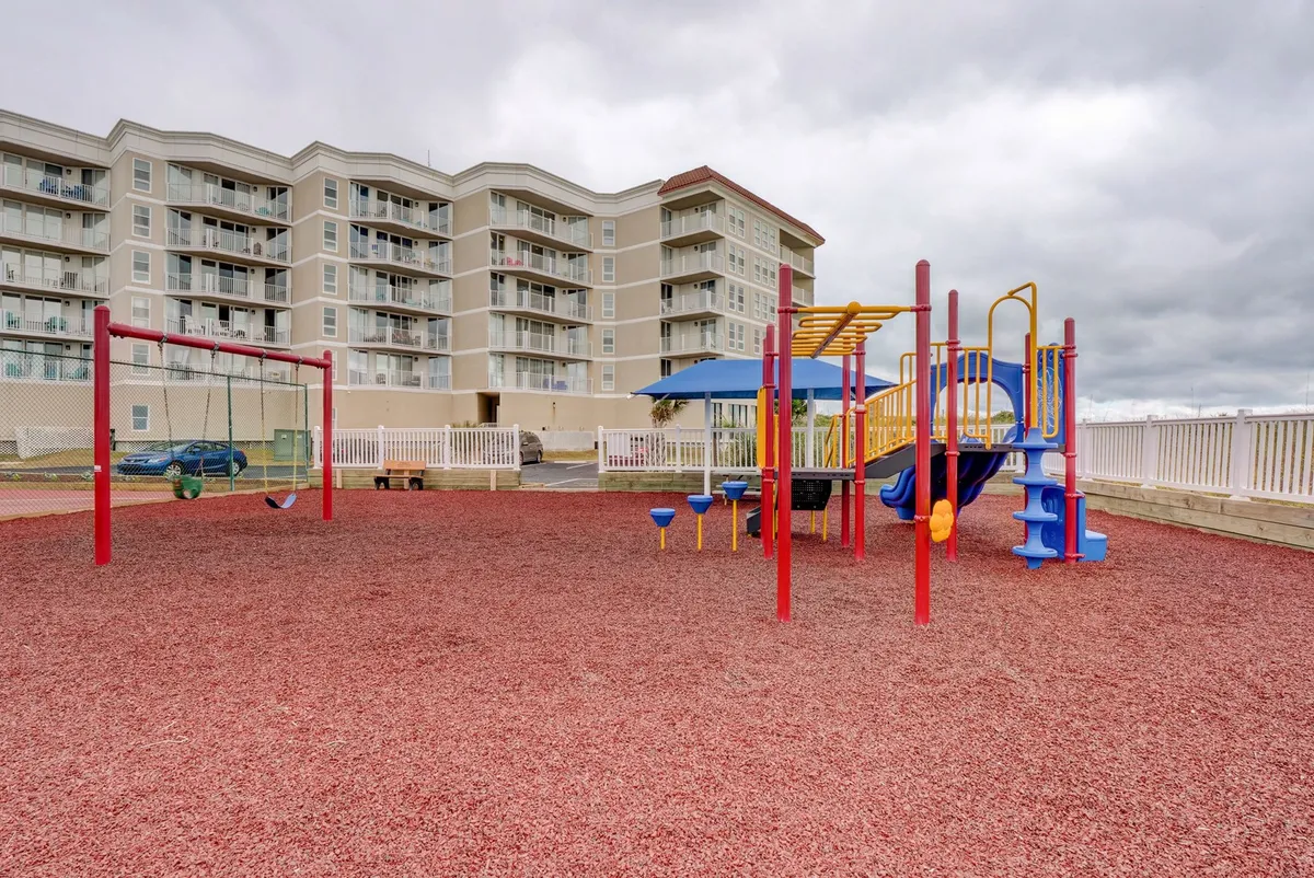 A lively playground scene with colorful equipment, swings, and children at play. Parents mingle nearby, and the soft red rubber ground ensures safety, while the apartment complex in the background signals the family-oriented environment.