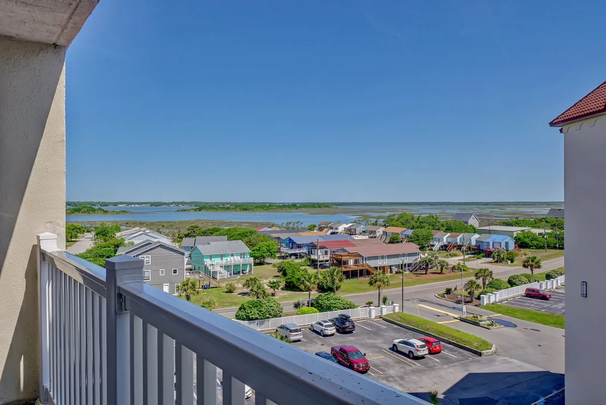 A tranquil waterfront view featuring colorful houses, a road, and a parking area, as seen from a vantage point ideal for balcony relaxation. The peaceful mood suggests quiet mornings or evenings spent together, with opportunities to explore the scenic locale nearby.