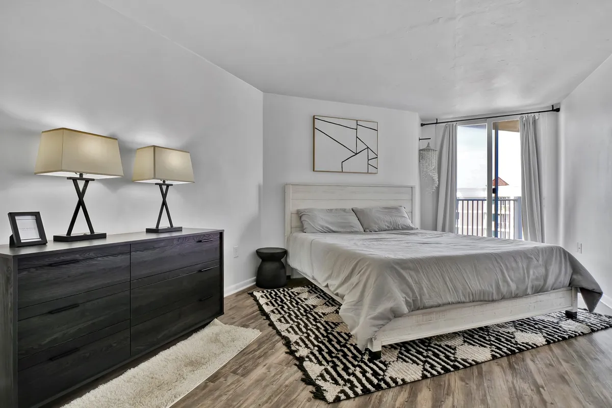 A cozy bedroom with a comfortable bed, elegant dressers, table lamps, and wall art. Ample natural light streams in through a window, creating a welcoming and tranquil environment ideal for relaxation or reading.