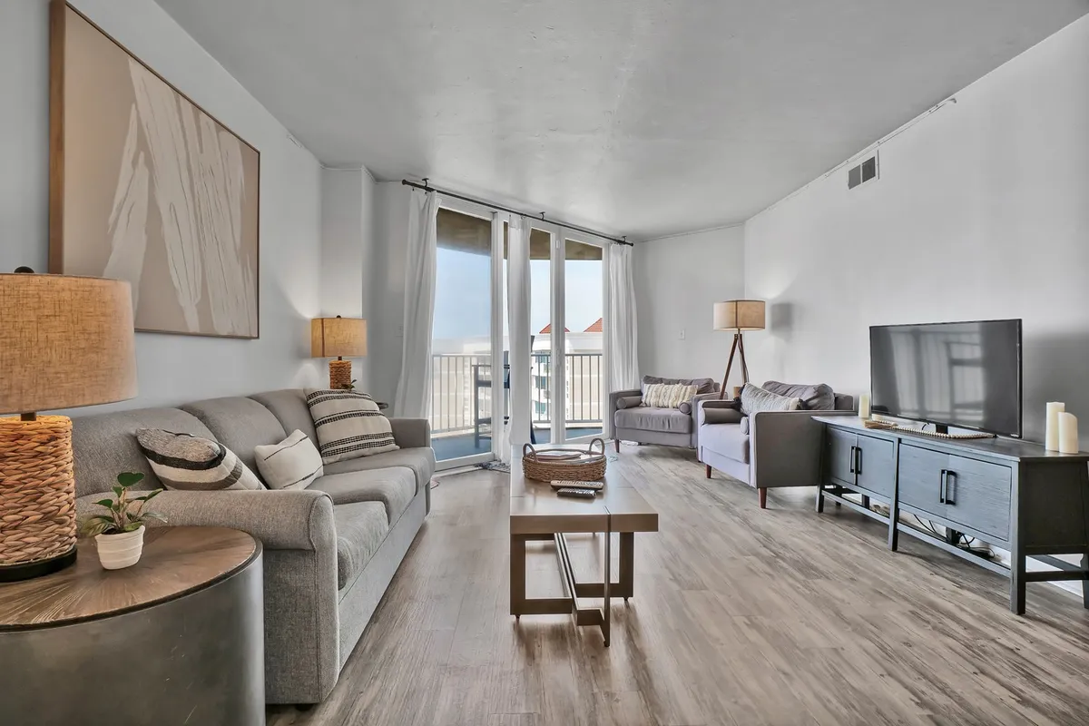 A spacious, comfortable living room featuring a sofa, lamps, coffee table, television, and sliding glass doors that open out to a balcony. The room is filled with morning sunlight and offers inviting spots for relaxation or family time.