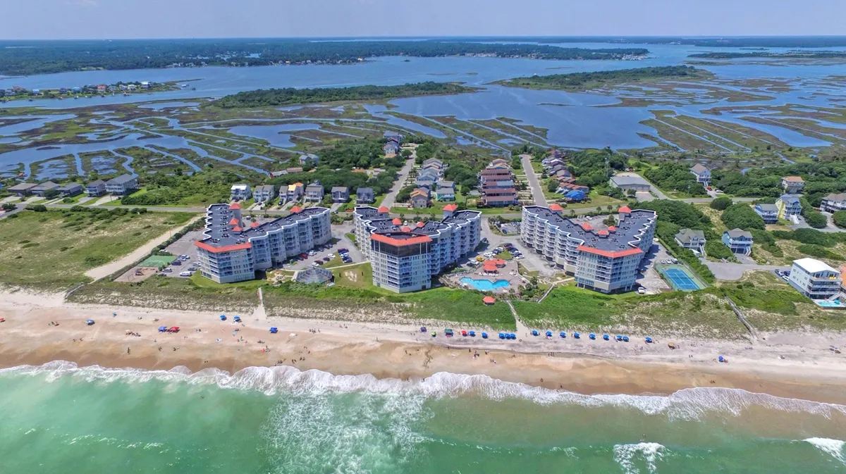 A vibrant resort pool scene at the edge of the beachfront, complete with colorful umbrellas, sunbathers, and the backdrop of marshlands and the Atlantic Ocean. The lively atmosphere invites guests to lounge, swim, and socialize in a quintessential vacation setting.