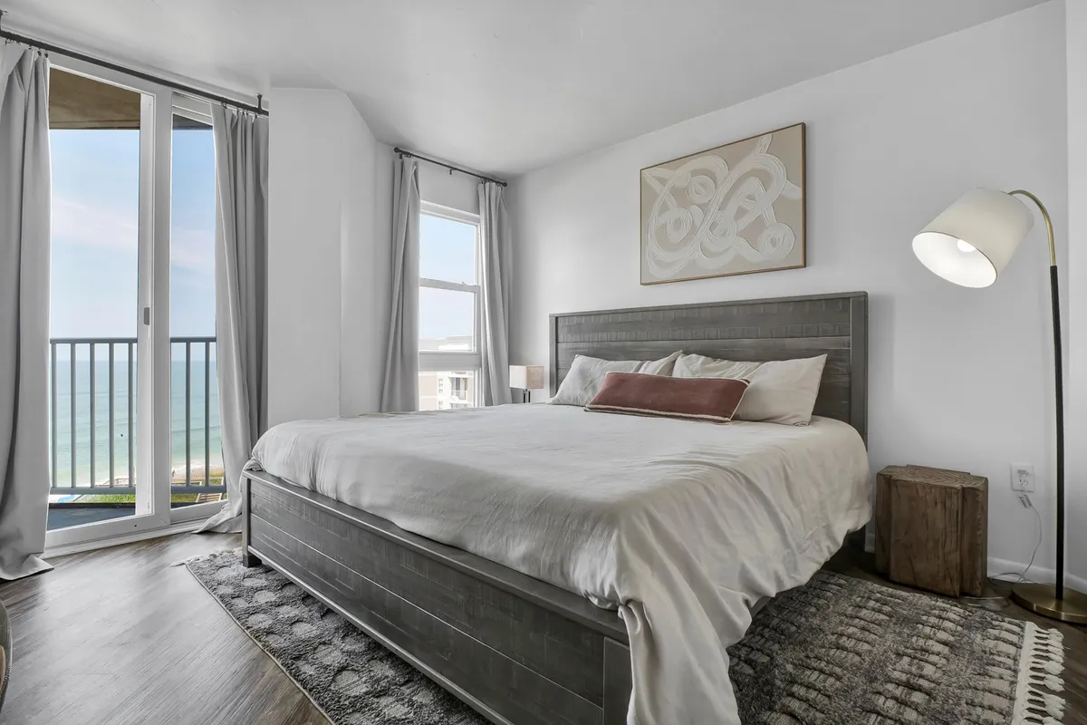 A serene bedroom with a large bed facing windows that overlook the ocean. Soft, coastal-themed decor, a reading lamp, and calming artwork create a tranquil atmosphere. Natural light floods the space, emphasizing the relaxing and restful qualities of the room.