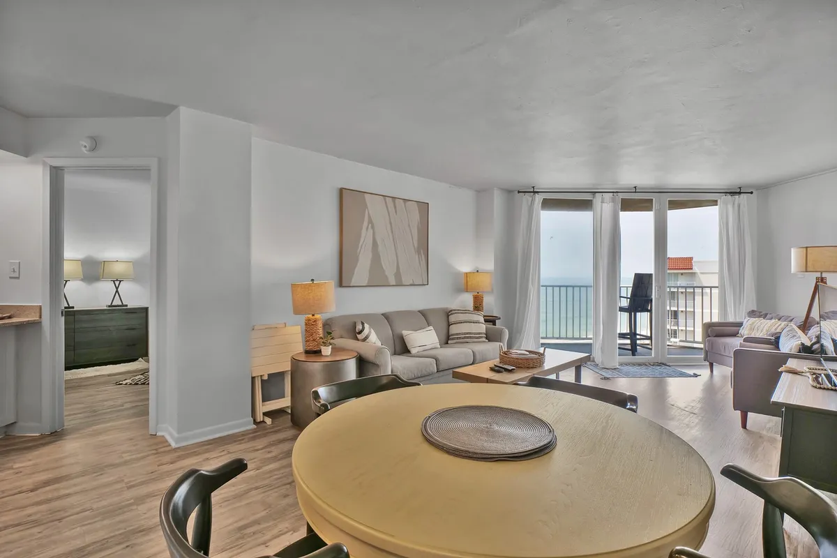 A cheerful, comfortable living-dining space with a modern dining table, plush sofa, and large windows offering panoramic ocean views. Artwork on the wall adds a touch of personality and warmth.