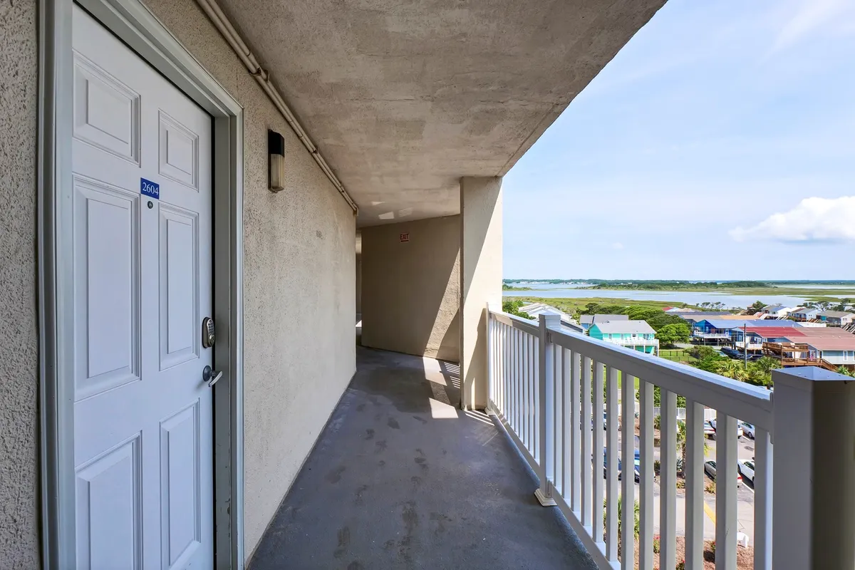 This image captures a peaceful exterior balcony scene with a white door, railing, and a scenic view of the coastline. The setting basks in gentle sunlight, suggesting the perfect spot for morning coffee or evening relaxation while gazing at the ocean.