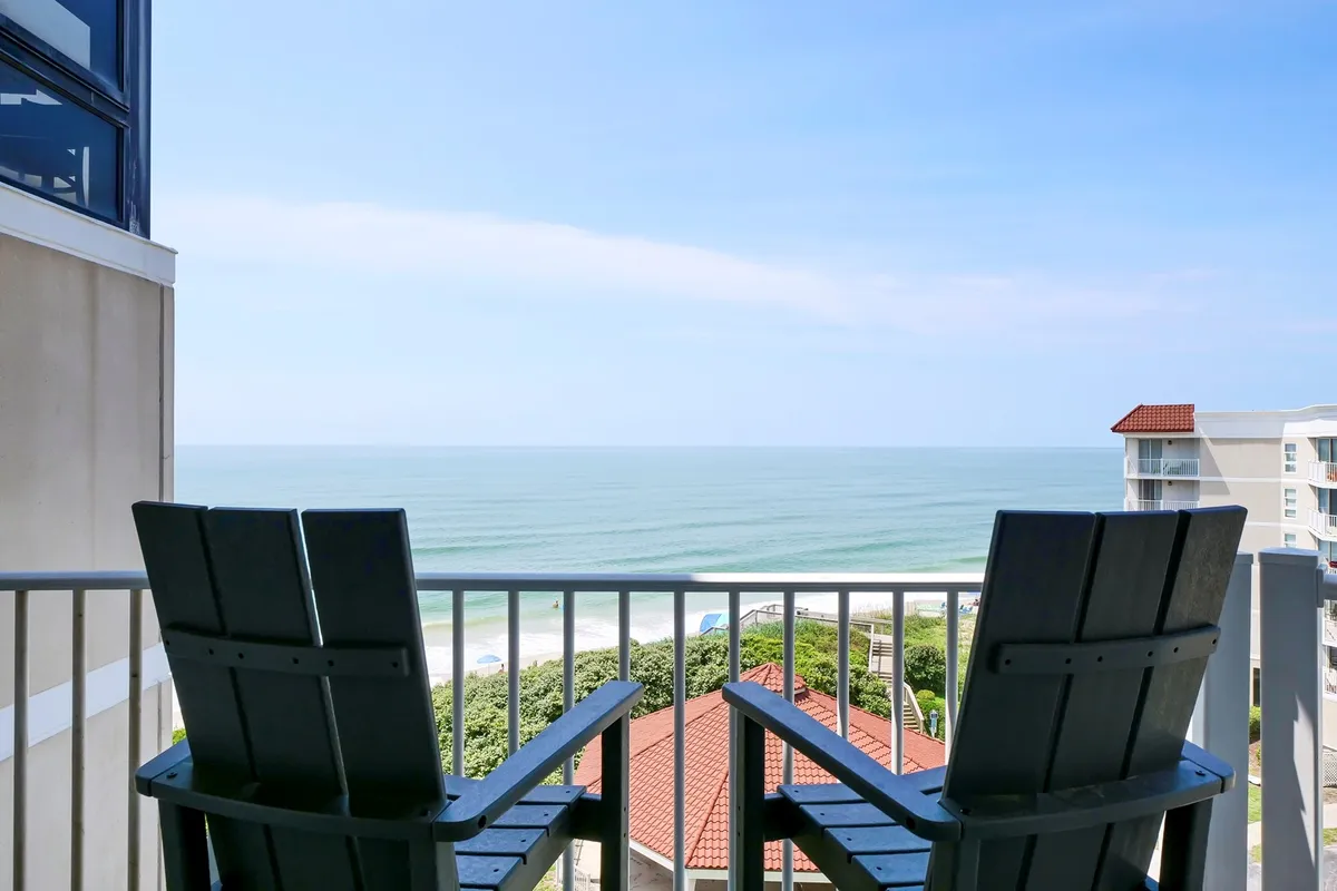 The image shows two inviting Adirondack chairs positioned on a private balcony, overlooking a breathtaking ocean view. Below, colorful beach umbrellas dot the sandy shore, suggesting a lively beach scene. The tranquil setting hints at moments of relaxation, whether watching the sunrise, enjoying a quiet read, or sharing conversation with a loved one while waves roll in below.