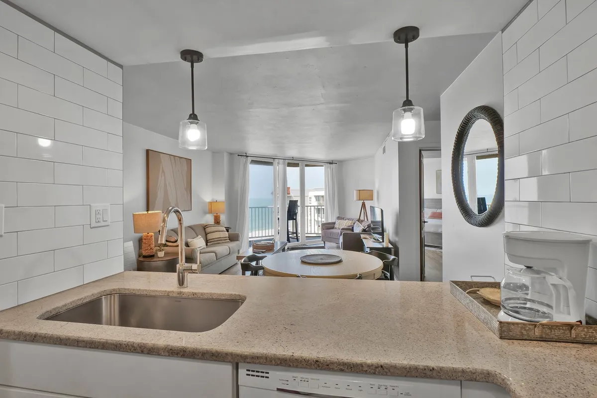 An open-plan living space featuring a kitchen island, large windows flooding the area with natural light, a comfortable seating area with sofas, and a round dining table. The windows offer a scenic ocean view, tying the interior to the coastal setting.